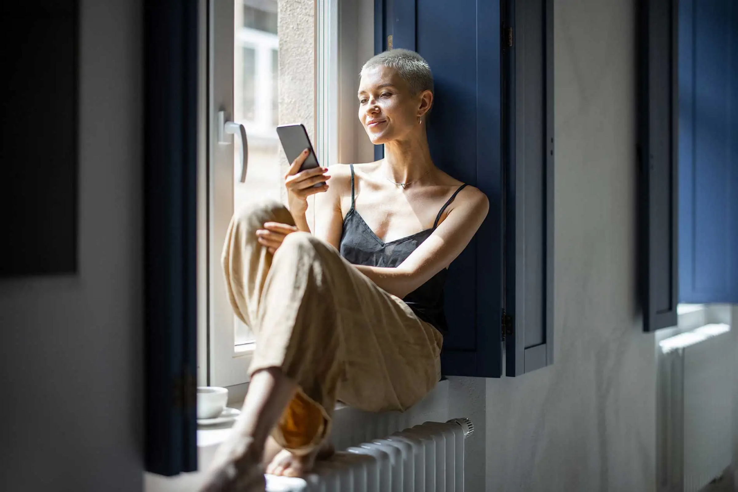 Brilliant Teaser Eine Frau sitzt auf einem Fenstersims und schaut auf ein Handy. brillant-teaser-app-bg.webp